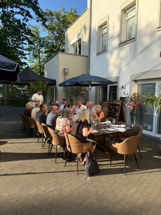 Gäste an einer langen Tafel vor der Vino Vista im Freien, unter einem Sonnenschirm im Abendlicht