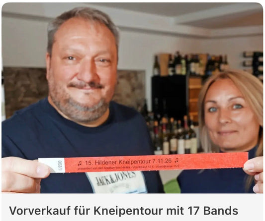 Der Organisator der Hildener Kneipentour und eine Inhaberin der Vino Vista Weinbar halten ein rotes Bändchen mit der Aufschrift „15. Hildener Kneipentour 7.11.26“ in die Kamera, dahinter die Weinregale